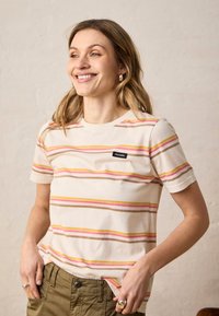 Femme souriante aux cheveux mi-longs portant un t-shirt crème à rayures horizontales roses, jaunes et marron, ainsi qu'un pantalon vert olive, mains dans les poches.