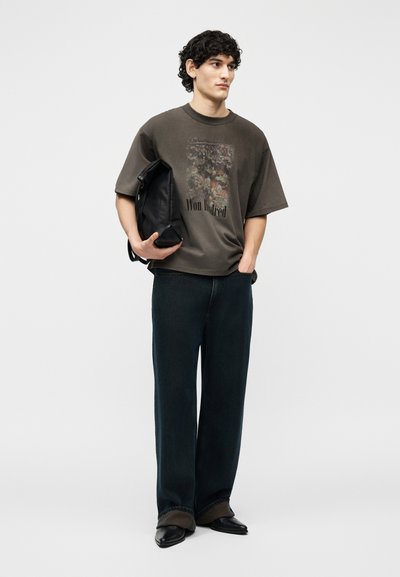 Νέος άνδρας με σγουρά μαλλιά στέκεται κρατώντας μαύρο σακίδιο πλάτης, φοράει σκούρο μπλουζάκι με σχέδιο, φαρδιά μαύρη παντελόνια και μαύρα παπούτσια μπροστά σε απλό φόντο.