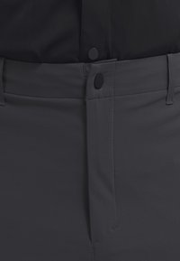 Primo piano di pantaloni grigio scuro con chiusura frontale a bottoni e passanti per cintura indossati con una camicia nera.