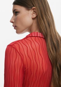 Rote durchsichtige Bluse mit einem strukturierten wellenförmigen Muster, einem spitzen Kragen und einer taillierten Silhouette mit langen Ärmeln, die ein glattes Rückenprofil zeigt.