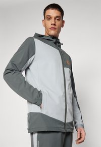 Helehall ja tumedhall zip-up jakk kapuutsiga, millel on geomeetriline muster, elastsed mansetid ja oranž logo rinnal.