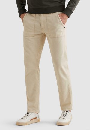 Mężczyzna noszący beżowe spodnie o prostych nogawkach z przednimi naszywanymi kieszeniami oraz białe trampki, ręce w kieszeniach, skrócone w pasie i przy kostkach.