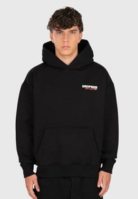 Jeune homme aux cheveux bouclés portant un hoodie noir avec une poche avant et le logo "DROPSIZE No Limits" sur la poitrine, se tenant devant un fond uni.