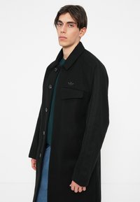 Cappotto in misto lana nero con chiusura a bottone, colletto e tasca sul petto. Presenta dettagli a coste sulle spalle e sulle maniche.