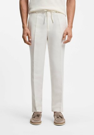 Personne portant un pantalon sur mesure écru avec une taille à cordon et des mocassins à pampilles beiges, debout sur un fond blanc.
