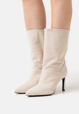 Bottines en daim beige avec un bout effilé, un tige ample et de fins talons hauts, présentant des détails de couture minimaux.