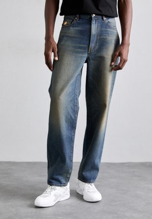 Blå denimjeans med en lös passform, med urtvättade områden, vanliga fickor och knappgylf. Matchas med vita sportskor.