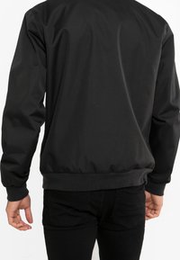 Sort bomberjakke med en glat, mat overflade. Har ribbede manchetter og kant, afslappet pasform og minimalistisk design uden udsmykninger.