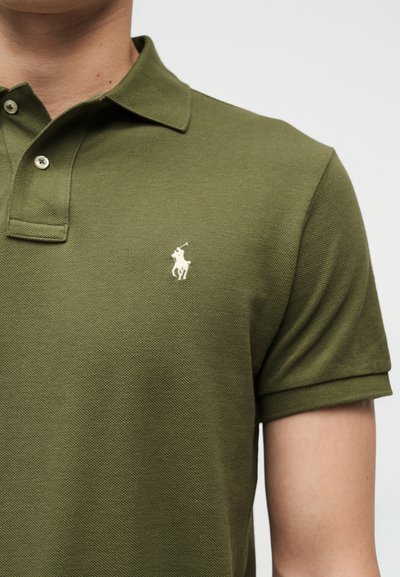 Άνδρας που φοράει πόλο μπλούζα σε ελιά πράσινο χρώμα με κοντά μανίκια, με κουμπωτό γιακά και μικρό λευκό κεντημένο λογότυπο αλόγου και αναβάτη στο στήθος.