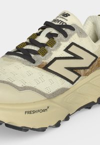 Κοντινή λήψη μπεζ παπουτσιού New Balance για τρέξιμο με μαύρα και ελαιοπράσινα κορδόνια, μεγάλο λογότυπο «N» και κείμενο «Fresh Foam X» στη σόλα.