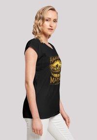 Camiseta negra de manga corta con un gráfico amarillo que dice "Hakuna Matata" de El Rey León. Tejido suave, corte holgado, diseño con hombreras.