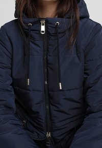 Chaqueta acolchada azul marino con cierre frontal, capucha ajustable con cordón y detalles metálicos. Textura suave con un diseño elegante.