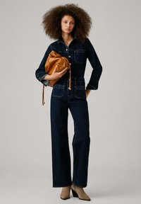 Denim jumpsuit i mörk indigo med knappstängning, bröstfickor och vida ben. Modellen håller en tanfärgad läderdragsväska. Beiga ankelskor.
