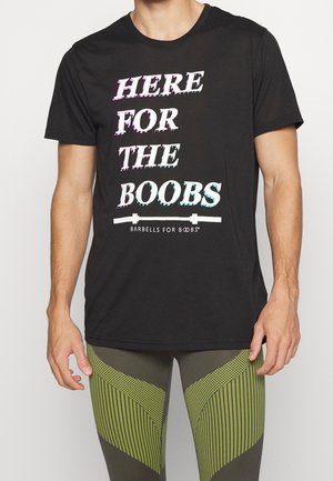 Mężczyzna w czarnym T-shircie z białym i kolorowym napisem "Here for the Boobs" oraz w zielonych legginsach sportowych z wzorem.