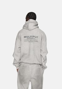 Sivá mikina s kapucňou a zodpovedajúcimi teplákmi; látka vyzerá mäkká s česanou textúrou. Na zadnej strane je logo "MULTIPLY APPAREL" v tmavosivej farbe.