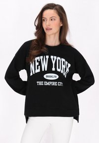 Fekete pulóver hosszú ujjakkal, feltűnő fehér szöveggel: "NEW YORK BROOKLYN A BIRODALOM VÁROSA." Kerek nyakú, laza fazonú, oldalhasítékokkal.
