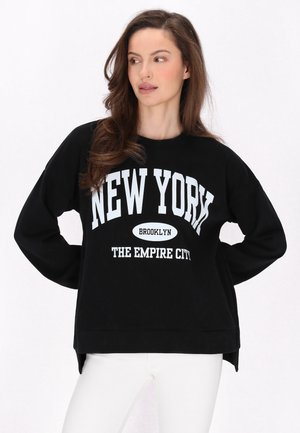 Fekete pulóver hosszú ujjakkal, feltűnő fehér szöveggel: "NEW YORK BROOKLYN A BIRODALOM VÁROSA." Kerek nyakú, laza fazonú, oldalhasítékokkal.