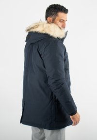 Manteau d'hiver bleu marine rembourré avec une capuche en fourrure, poche zippée sur la poitrine, coupe décontractée et poches latérales. Texture lisse et isolation chaude.