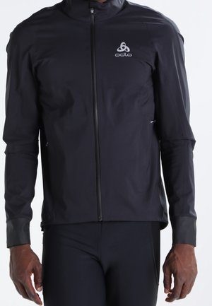 Veste de sport noire à fermeture éclair intégrale avec logo Odlo sur la poitrine, portée par une personne à la peau foncée, assortie à un pantalon de sport noir.