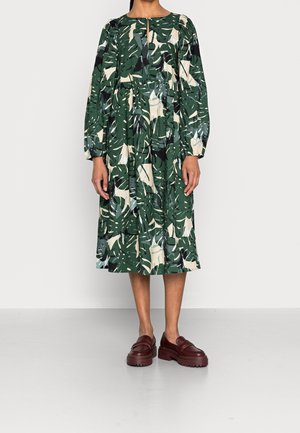 Vrouw die een midi-jurk met lange mouwen draagt, bedrukt met groene, beige en zwarte bladeren, en bruine chunky loafers, staand op een grijze vloer.