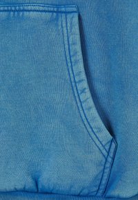 Sweat-shirt bleu avec poche kangourou, tissu texturé, coutures détaillées et ourlet côtelé. Le matériau a un aspect doux et usé.