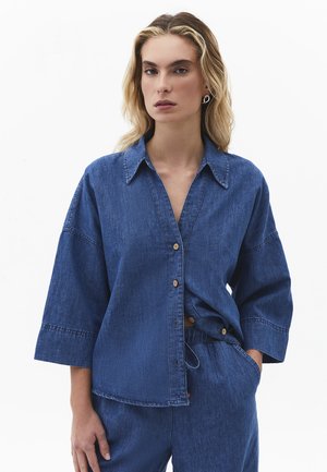 Kvinde med blondt hår iført løs knappet denimskjorte og matchende denimbukser, stående foran en ensfarvet hvid baggrund.