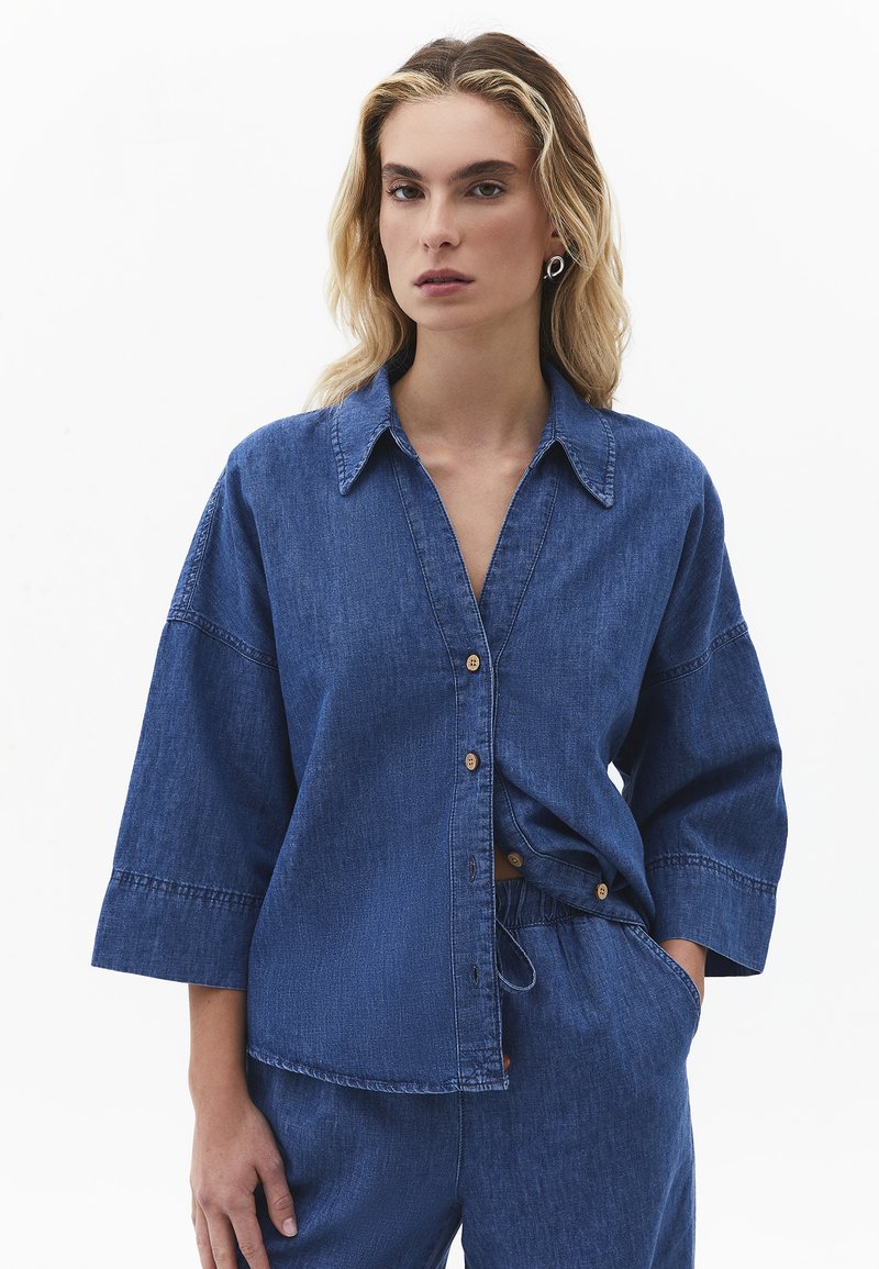 Kvinde med blondt hår iført løs knappet denimskjorte og matchende denimbukser, stående foran en ensfarvet hvid baggrund.