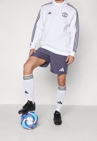Adidasin valkoinen huppari purppuranvärisin korostuksin, harmaat shortsit ja mustat jalkapallokset. Pelaajan jalka lepää monivärisellä jalkapallolla.
