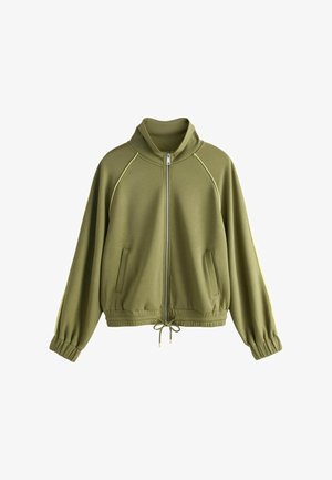 Giovane donna con lunghe trecce che indossa una giacca verde oliva con zip e pantaloni coordinati, mani nelle tasche, sfondo neutro.
