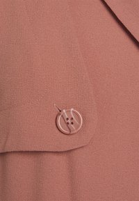 Tejido de color rosa con un acabado texturizado, que presenta un botón redondo con rayas y un detalle de costura simple en la solapa.
