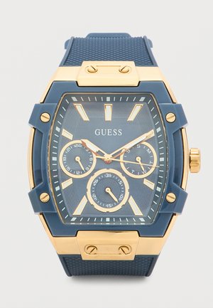 Μπλε και χρυσό ρολόι χειρός Guess με ορθογώνιο καντράν, τρεις υποκαντράν, χρυσούς δείκτες ωρών και υφασμένο μπλε λουράκι.