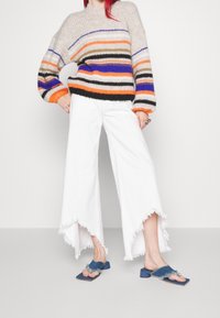 Pull en tricot à rayures multicolores orange, bleu et vert. Pantalon blanc à jambes larges avec des ourlets effilochés et sandales bleues à bouts ouverts.