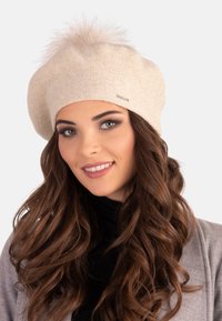 Chapeau béret tricoté beige avec un doux pompon en fausse fourrure sur le dessus et un détail de logo discret. Texture lisse et forme arrondie.