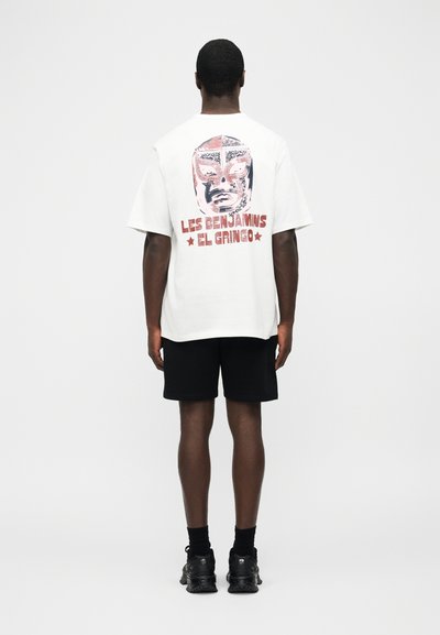 Άνδρας με λευκό φαρδύ T-shirt με κόκκινη και μαύρη γραφική μάσκα lucha libre και κείμενο "Les Benjamins El Gringo" στην πλάτη, μαύρη βερμούδα, μαύρα παπούτσια.