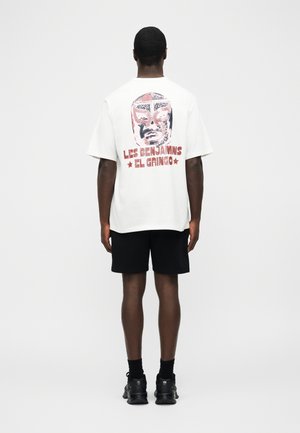 Άνδρας με λευκό φαρδύ T-shirt με κόκκινη και μαύρη γραφική μάσκα lucha libre και κείμενο "Les Benjamins El Gringo" στην πλάτη, μαύρη βερμούδα, μαύρα παπούτσια.