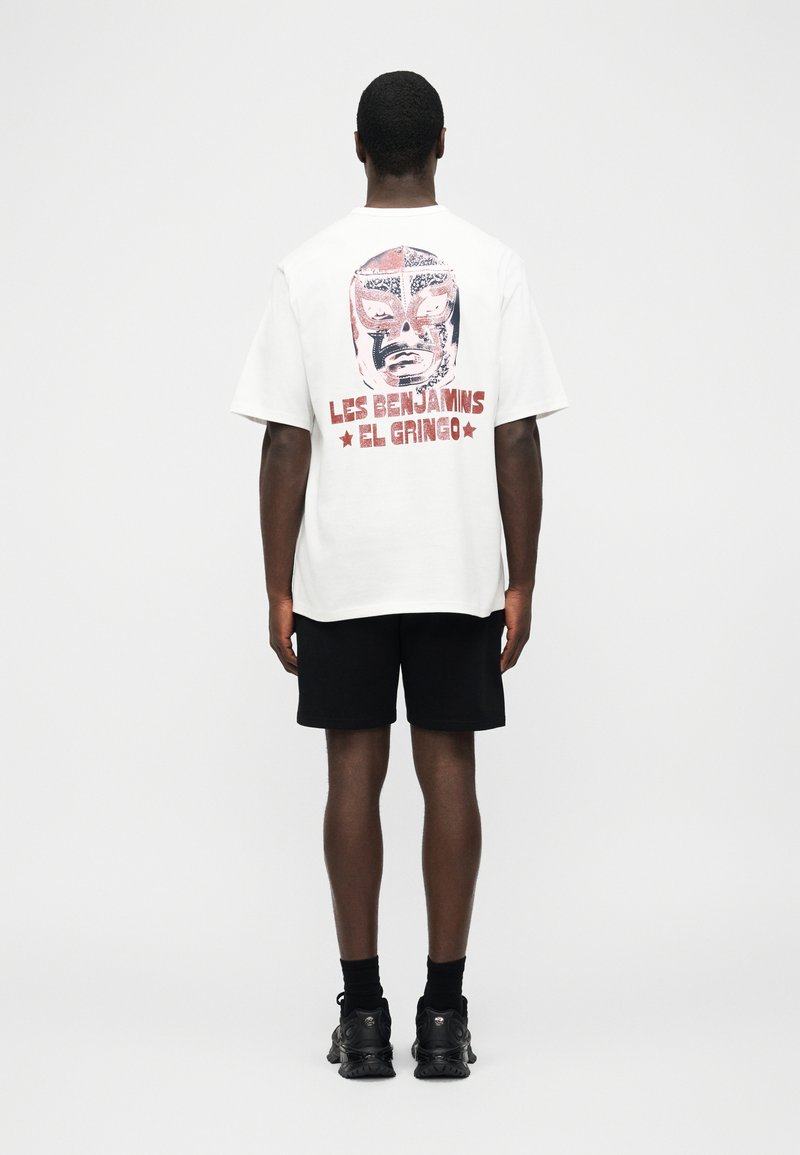 Mann trägt weißes, übergroßes T-Shirt mit rotem und schwarzem Lucha-Libre-Masken-Design und dem Text "Les Benjamins El Gringo" auf der Rückseite, schwarze Shorts, schwarze Schuhe.