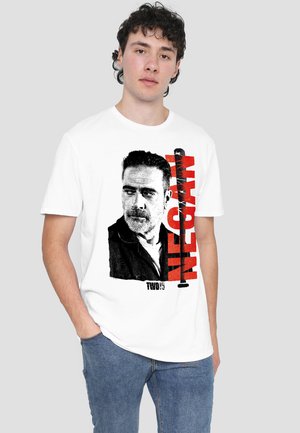 Young man with short curly hair wearing a white T-shirt featuring a black and white portrait and red text "NEGAN".