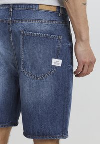 Shorts en denim bleu foncé, présentant une coupe droite, des poches arrière et une étiquette tissée à la taille, avec une texture lisse.