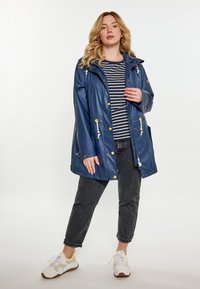 Tamsiai mėlynas vandeniui atsparus lietpaltis su kapišonu, geltonomis raišteliais, spaudėmis ir priekinėmis kišenėmis. Po juo vilkima juostuota marškinėliai ir džinsiniai kelnių.