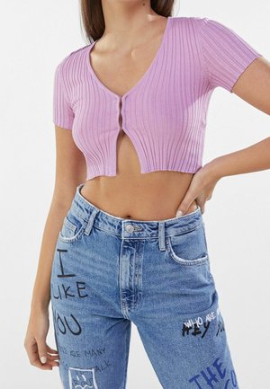 Crop top lilas clair côtelé avec un décolleté en V profond, des manches courtes et une fermeture à bouton. Assorti à un jean bleu taille haute avec un texte graphique.