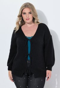 Svart cardigan med V-ringning, texturerat stickat material, överdimensionerade ärmar och knytningar framtill och i nederkant. Bärs över en teal-färgad topp.