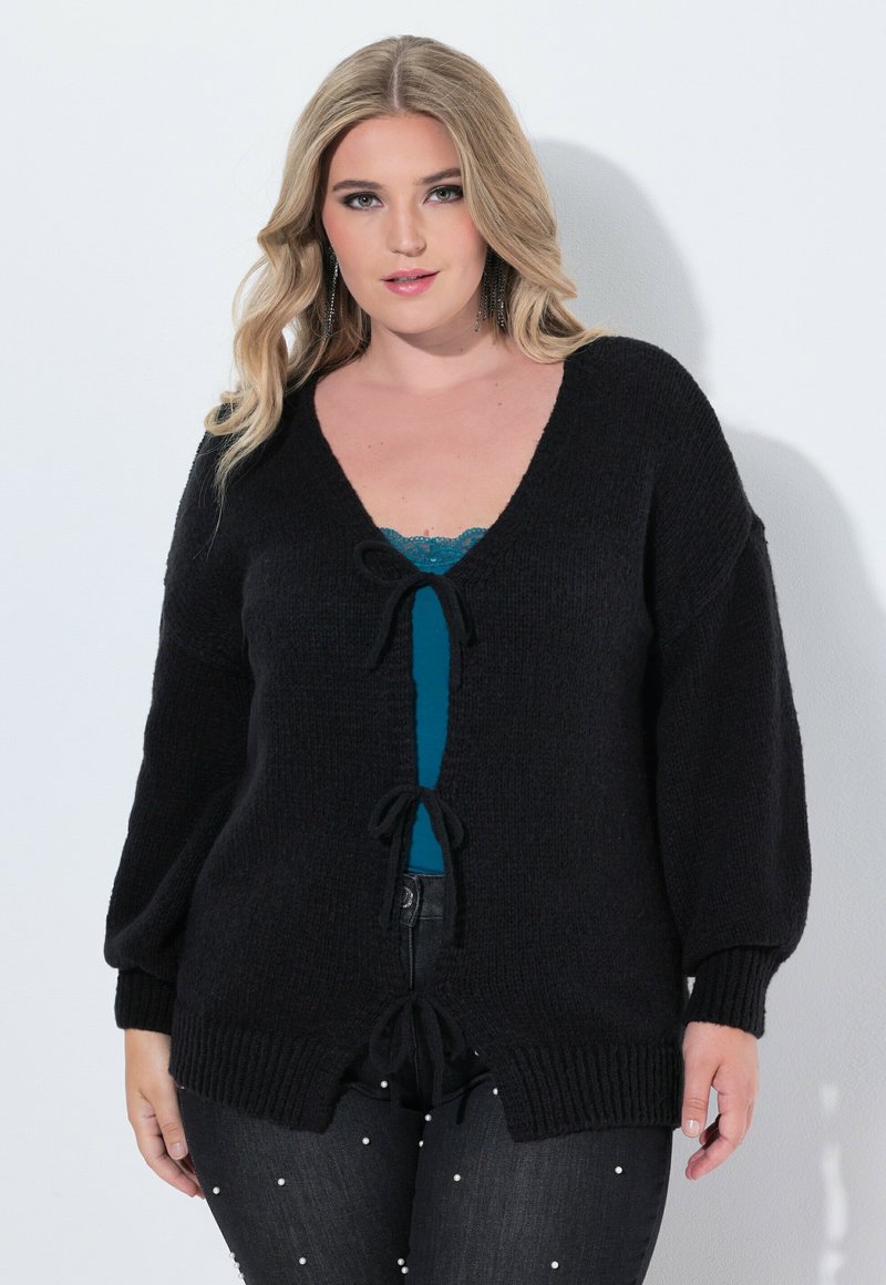 Svart cardigan med V-ringning, texturerat stickat material, överdimensionerade ärmar och knytningar framtill och i nederkant. Bärs över en teal-färgad topp.