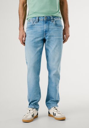 Mann trägt hellblaue Jeans mit geradem Bein und weiße Sneaker mit schwarzen Akzenten, steht vor einem einfarbigen Hintergrund.