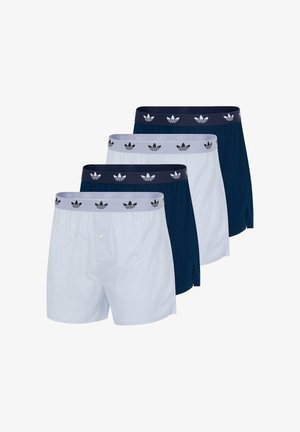 Giovane uomo in piedi a piedi nudi che indossa boxer blu navy con fascia elastica con marchio, su sfondo bianco uniforme.