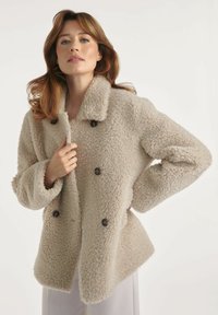 Kobieta w beżowym, puszystym, podwójnym płaszczu zapinanym na guziki, trzymająca kołnierz jedną ręką, stojąca na jednolitym jasnym tle.