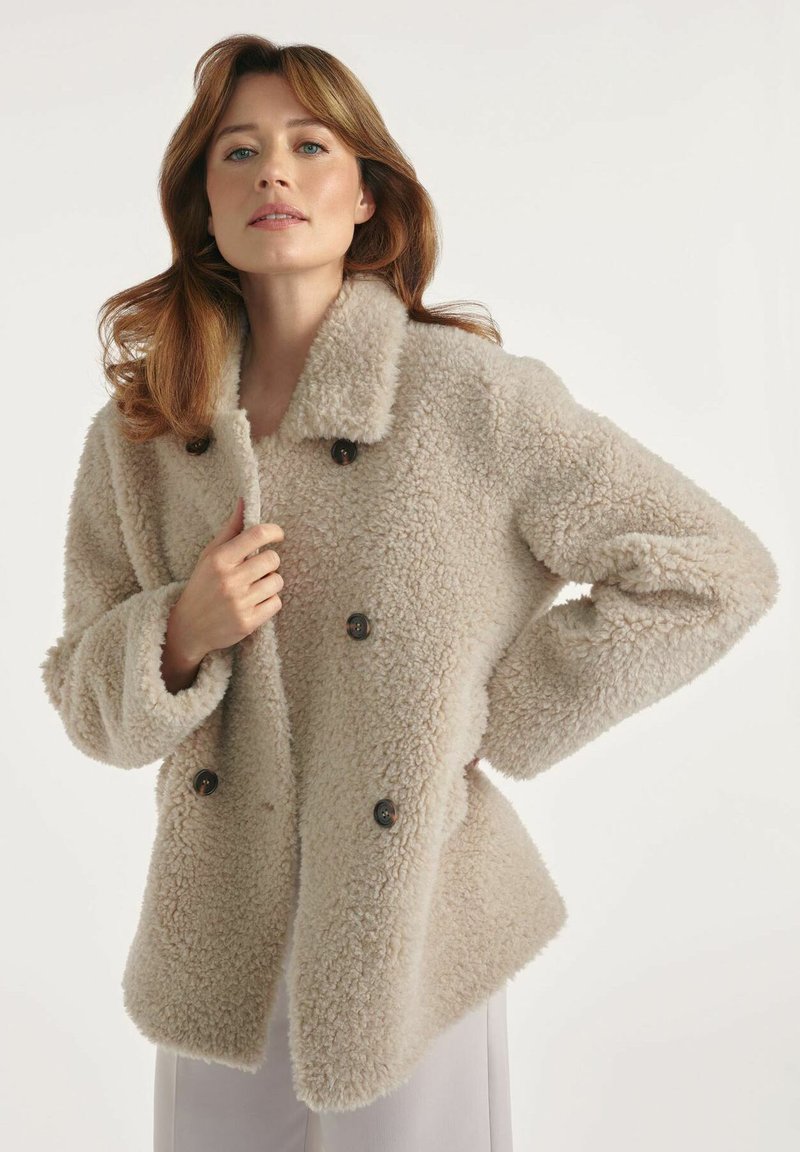 Kobieta w beżowym, puszystym, podwójnym płaszczu zapinanym na guziki, trzymająca kołnierz jedną ręką, stojąca na jednolitym jasnym tle.