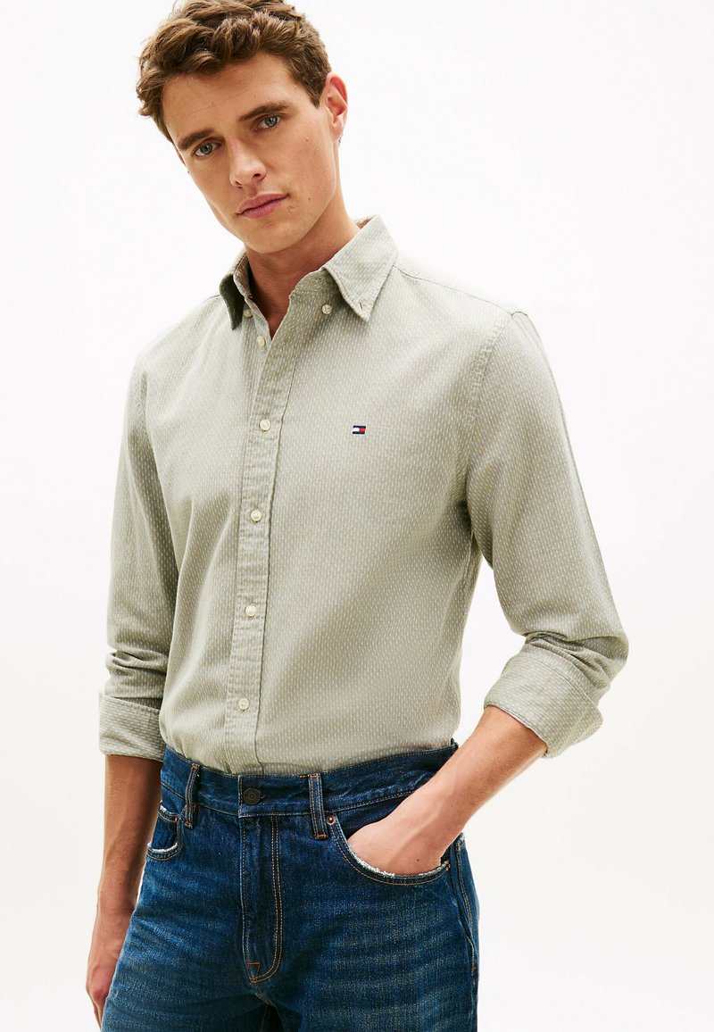 Homme portant une chemise à manches longues à motifs vert clair et un jean bleu, une main dans la poche, posant devant un fond uni.