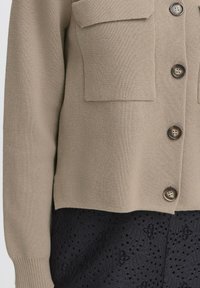 Cardigan a maglia beige con due tasche sul petto, tessuto a motivo scuro sotto e bottoni marroni. Tessuto texturizzato e forma strutturata.