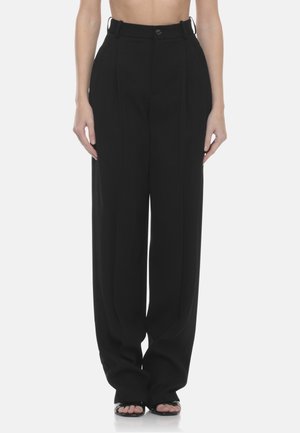 Donna che indossa pantaloni neri a vita alta, a gamba larga, con pieghe frontali e sandali neri aperti sulle dita, su uno sfondo semplice.