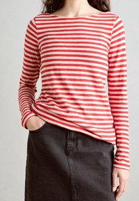 Rød- og cremegulstribet langærmet skjorte med bådhals, båret med en sort denimnederdel. Har en afslappet pasform og en hånd synlig i lommen.